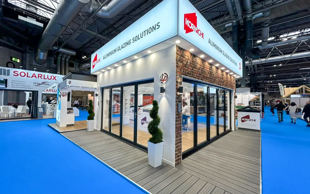 A wide shot of the National Homebuilding & Renovating Show floor before the show begins. The Korniche stand occupies a prominent location, showcasing bi-folding doors, roof lanterns, and a Flat Glass Rooflight System.
