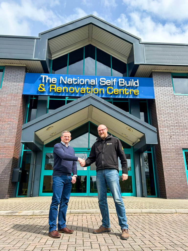an Bousfield, Korniche Marketing Manager, and Gareth Nicholas, NSBRC Business Development Manager, shaking hands in front of The National Self Build & Renovation Centre sign.