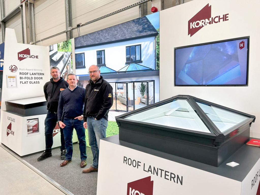 Three people at a Korniche trade show stand. From left to right are: a man in a Sevenday branded hoody (Area Manager from Sevenday Windows, Doors & Conservatories), a man in a jumper smiling (Ian Bousfield, Korniche Marketing Manager), and a man in a NSRBC branded fleece (Gareth Nicholas, NSBRC Business Development Manager)