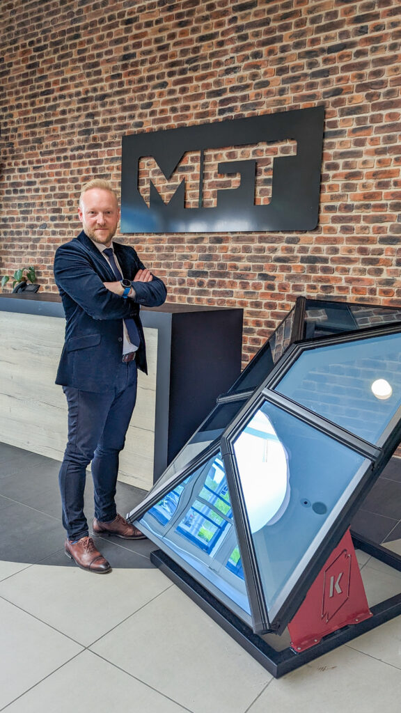 Bradley Gaunt, Managing Director of Made For Trade, in a suit with arms crossed, standing next to a Korniche Roof Lantern with the Made For Trade logo on a brick wall.