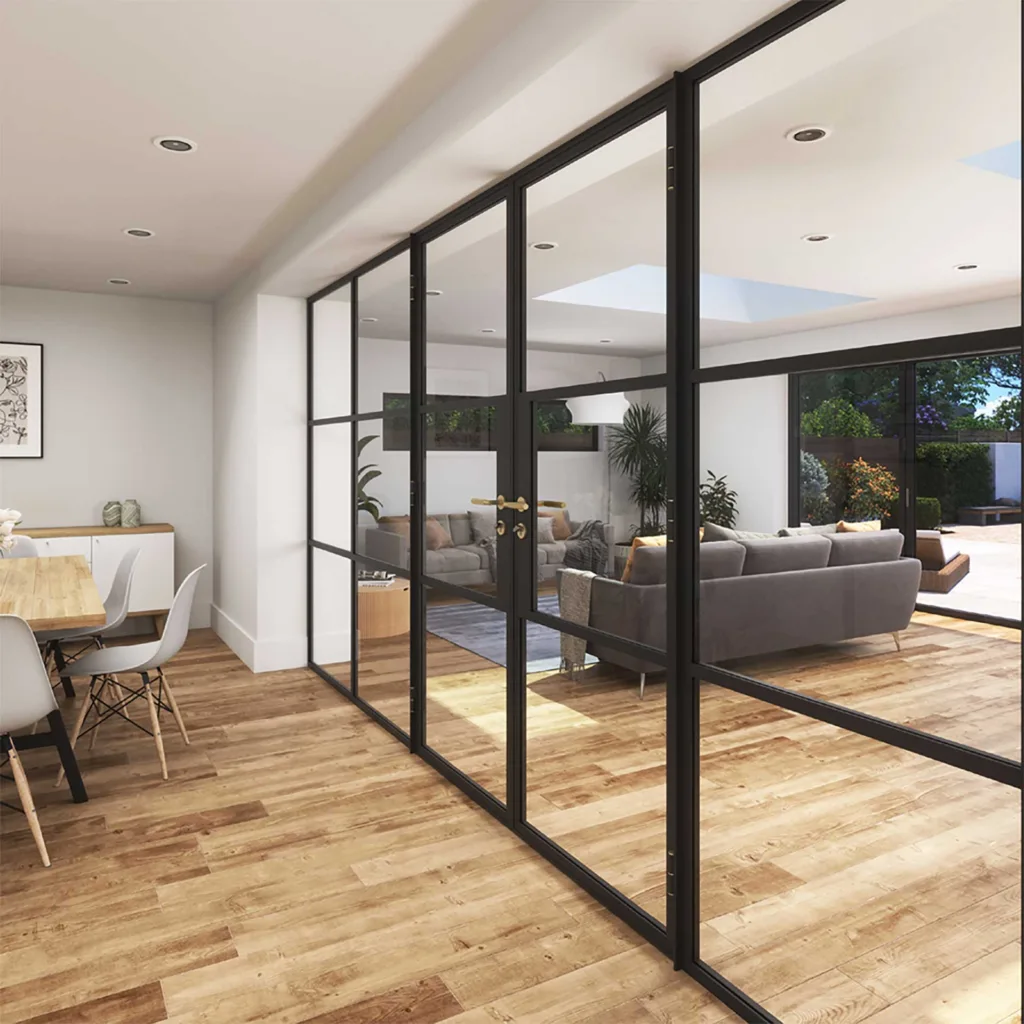 Korniche glass partition system with black frames dividing an open-plan living space. The glass panels allow light to flow between a living area with a grey sofa and rug, and a dining area with a wooden table and chairs, next to a modern kitchen with grey cabinets and a breakfast bar.