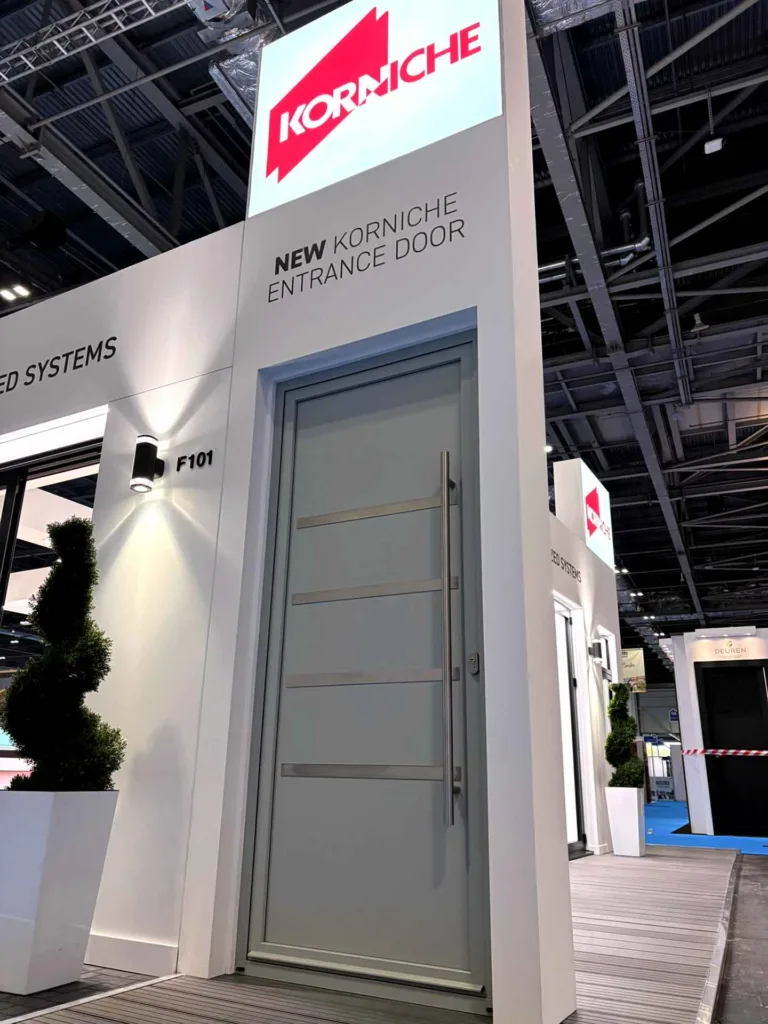Modern light grey Korniche aluminium entrance door with long vertical stainless steel pull handle on display at Stand F101, London Homebuilding & Renovating Show 2026.