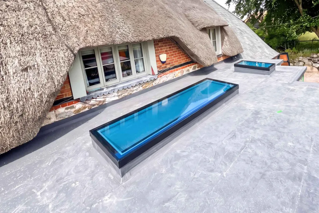 Two Korniche Flat Glass Rooflights with dark grey frames installed on a flat roof extension beside a traditional thatched cottage.
