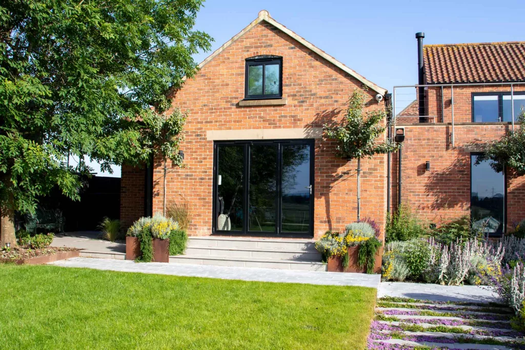 Black Korniche aluminium Bifold Doors installed in a red brick home, opening onto a landscaped garden and patio.