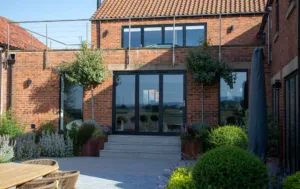 Dark grey Korniche aluminium Bifold Doors opening onto a raised patio, with a brick home and rooftop terrace above.