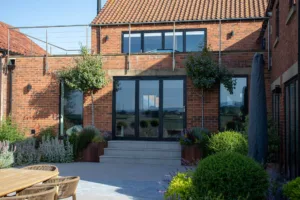 Dark grey Korniche aluminium Bifold Doors opening onto a raised patio, with a brick home and rooftop terrace above.