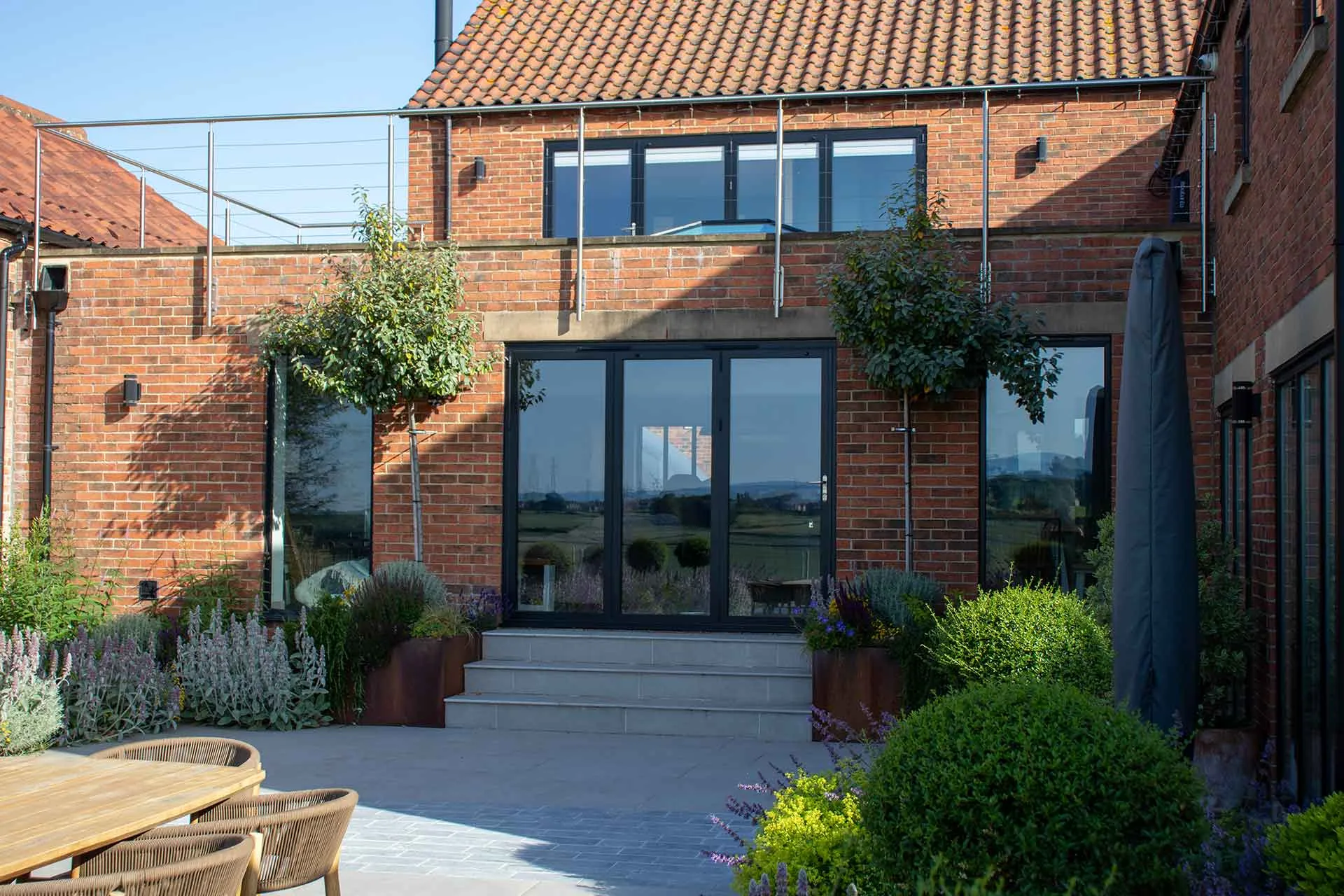 Dark grey Korniche aluminium Bifold Doors opening onto a raised patio, with a brick home and rooftop terrace above.