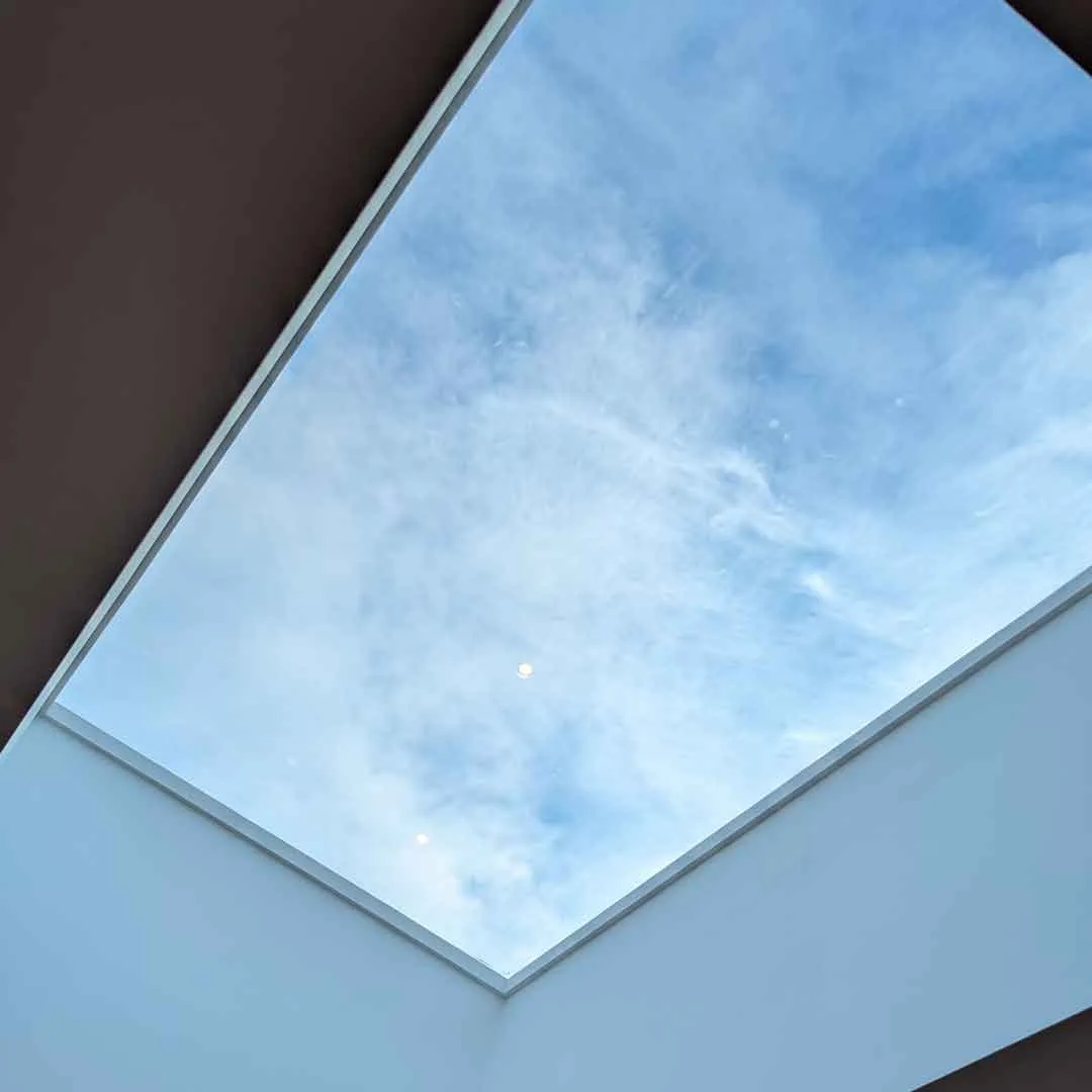 Interior view looking up through a Korniche Flat Glass Rooflight with a white internal finish, showing blue sky and clouds above.